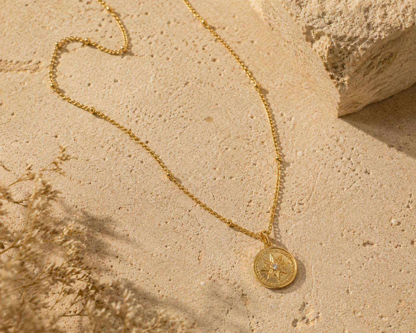 A delicate yellow gold necklace laid out on a textured, warm beige sand-like surface, creating a modern and earthy aesthetic